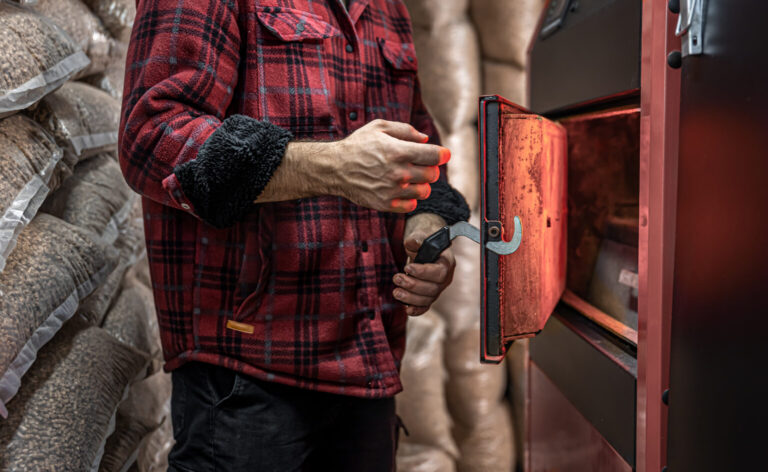 A person in a red plaid shirt opens the door of a red industrial machine, with bags of pellets stacked in the background.