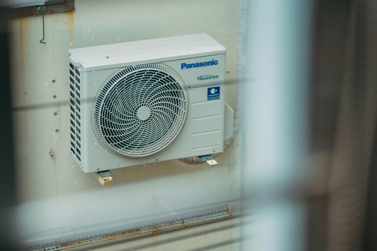 A white Panasonic inverter air conditioning unit is mounted on an exterior wall, viewed through a window with blurred bars in the foreground.