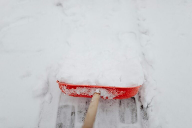 A close-up of a red snow shovel full of snow being used to clear a path on a snow-covered surface.