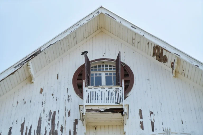 Weathered white wooden house with peeling paint, a round window with brown shutters, and a small balcony; a black bird perches on the railing.
