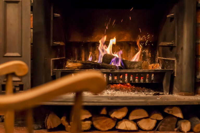 A fireplace with burning logs, emitting flames and sparks, with stacked firewood below and part of a wooden rocking chair visible in the foreground.