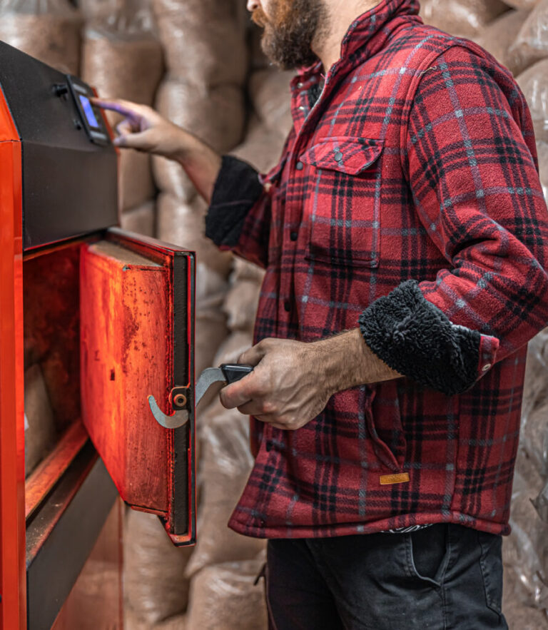 A man in a red plaid shirt operates a wood pellet boiler, adjusting controls with one hand and holding the boiler door open with the other.