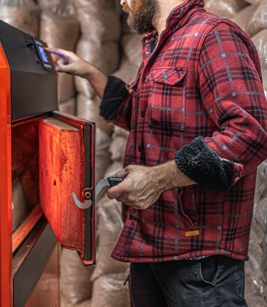 A man in a red plaid shirt operates a wood pellet boiler, adjusting controls with one hand and holding the boiler door open with the other.