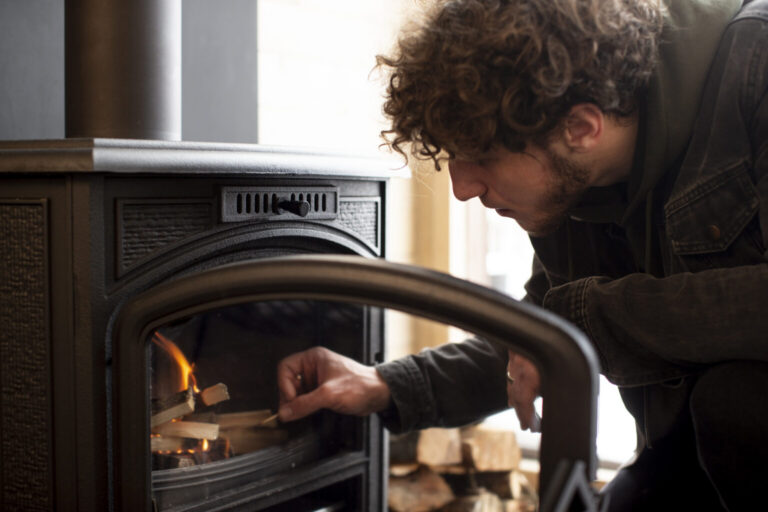 A person with curly hair is placing kindling into a wood stove, with a small fire already burning inside. Stacked firewood is visible in the background.