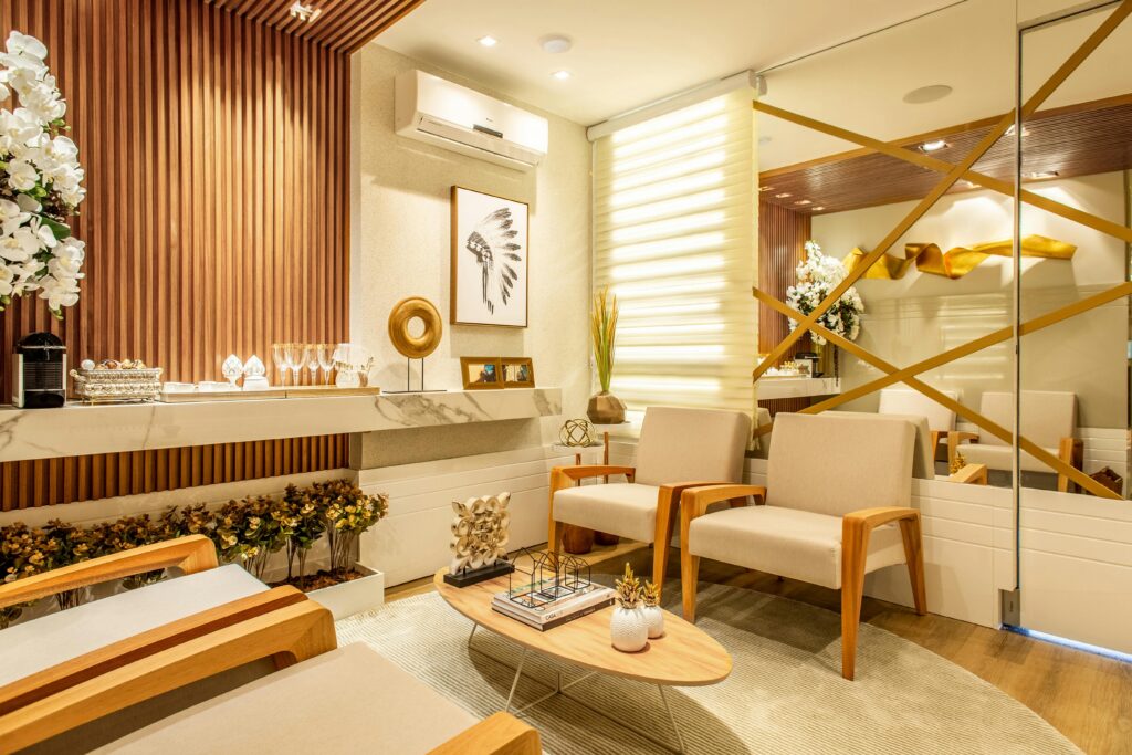 A modern waiting room with beige armchairs, a wooden coffee table, wall art, decorative plants, and a mirrored wall with gold accents.