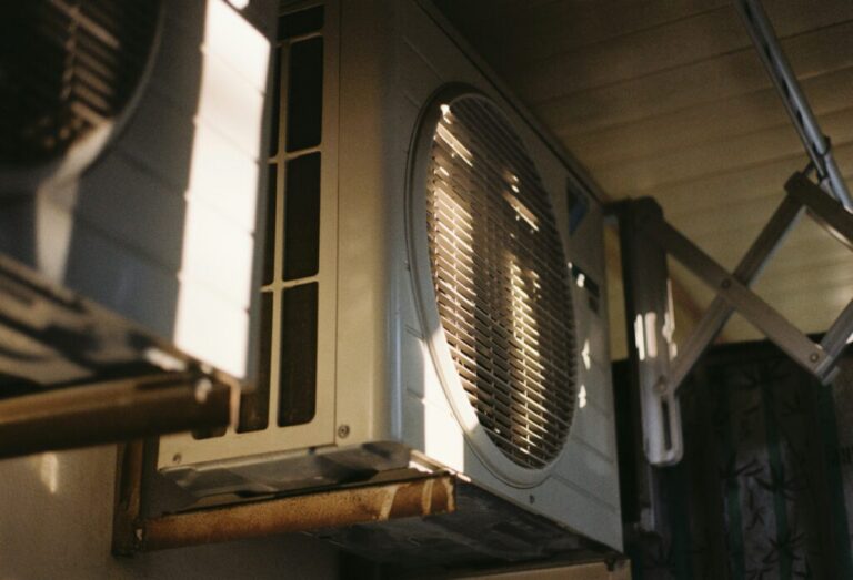 Close-up view of an outdoor air conditioning unit mounted on a wall, partially illuminated by sunlight.