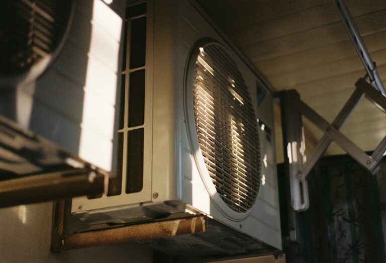 Close-up view of an outdoor air conditioning unit mounted on a wall, partially illuminated by sunlight.