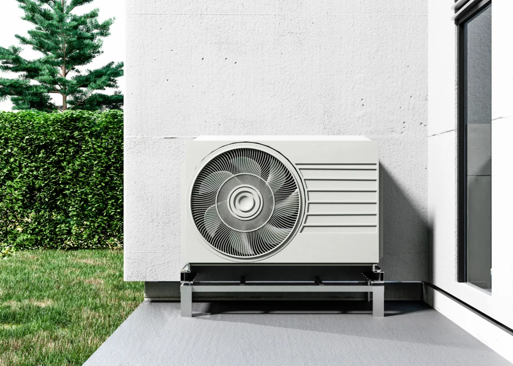 A heat pump unit is mounted on a metal stand outside a modern building, next to a green lawn and hedge.